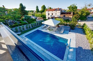 Una piscina rettangolare con bordo bianco, circondata da una zona prendisole con ciottoli. Si vedono lettini e un ombrellone bianco sotto un albero.