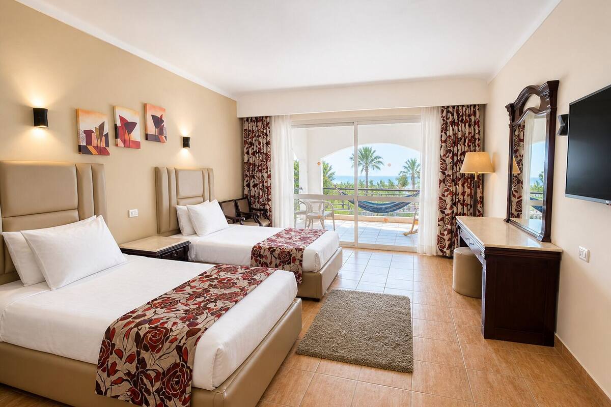 Habitación de hotel con dos camas, televisión, escritorio y balcón con vistas a palmeras y al mar.