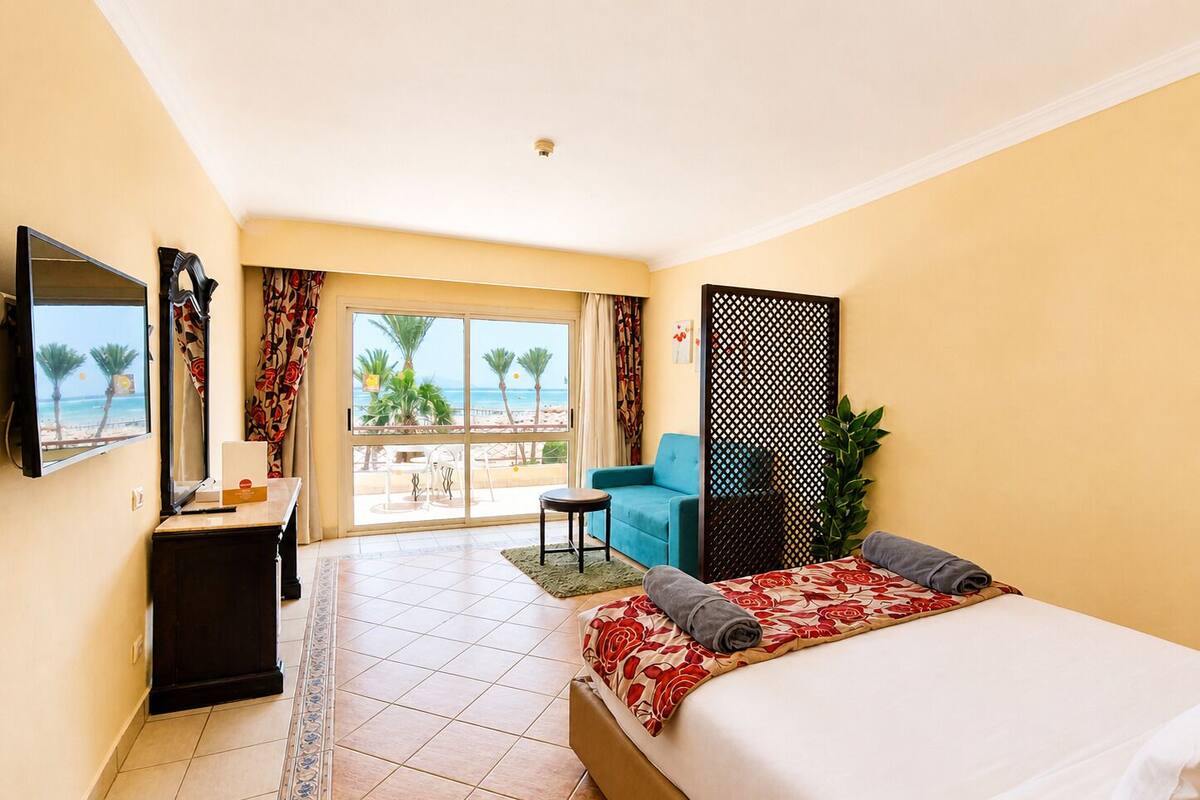 Habitación de hotel con cama, escritorio, silla, televisor, balcón con vista a la playa y una planta.