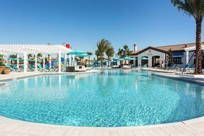 A large swimming pool with lounge chairs and umbrellas, surrounded by palm trees and a building with a tiled roof. - Family-Friendly Villa w Pool, Hot Tub & Game Room (Kissimmee)