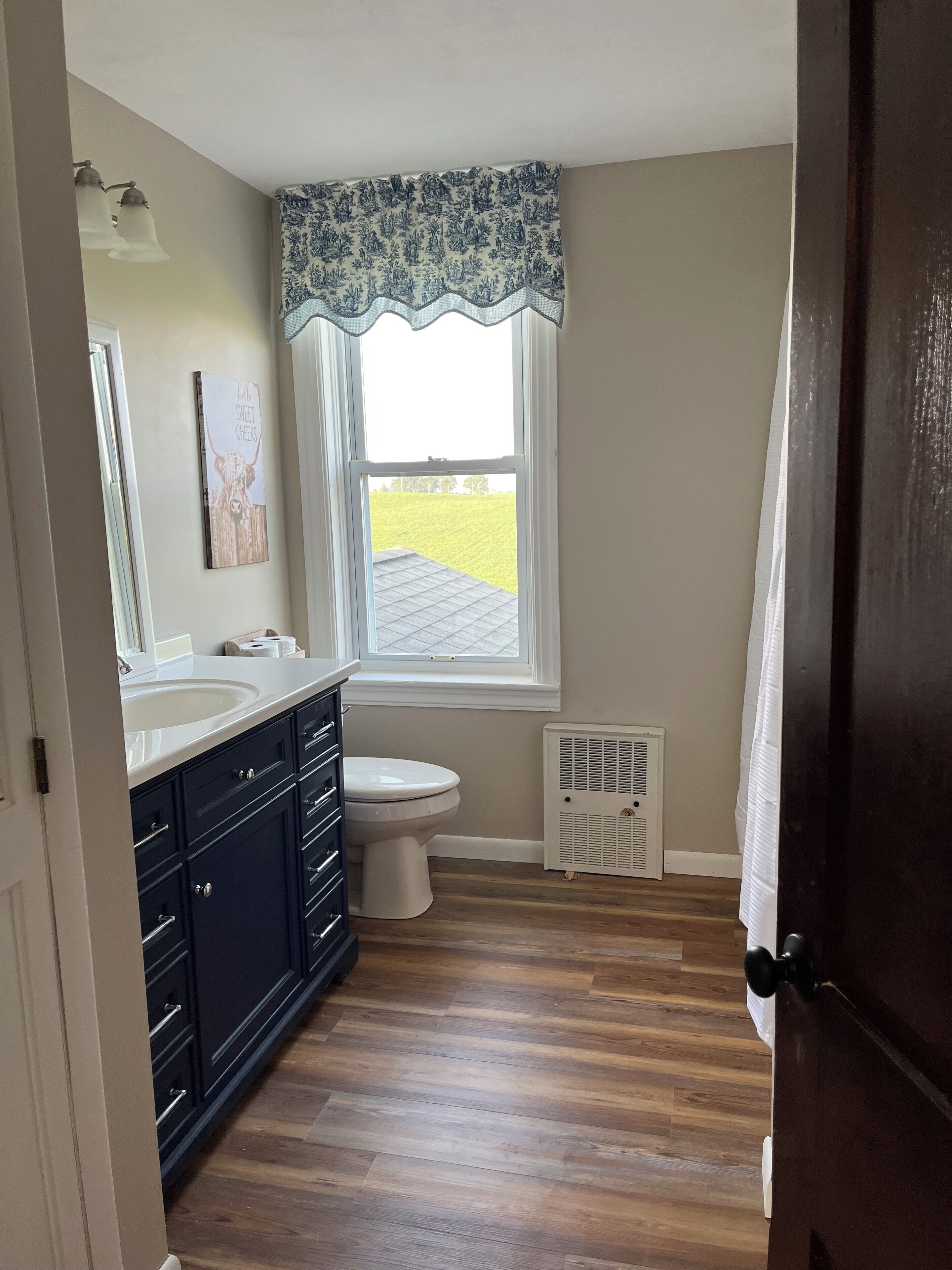 A bathroom with a double sink, dark cabinets, a toilet, and a window with a view of a green field.