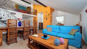 A living room with a blue sofa, wooden chairs, a coffee table, and a staircase with a colorful railing. -  De Vivo Realty - La Mela Apartment (Meta)