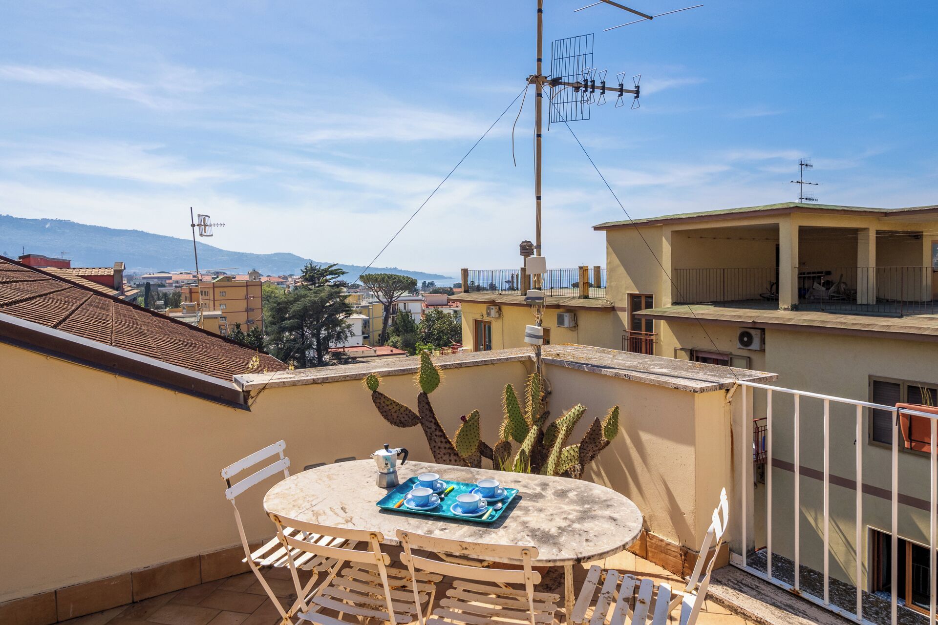 Eine Dachterrasse mit Tisch für vier Personen, umgeben von Kakteen, mit Blick auf eine Stadtlandschaft mit Gebäuden und einem fernen Berg.
