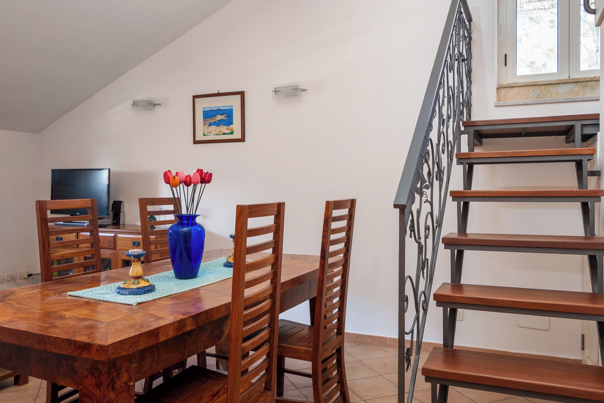 Un comedor con una mesa de madera, sillas, un jarrón azul con flores, un cuadro en la pared, un televisor y una escalera.