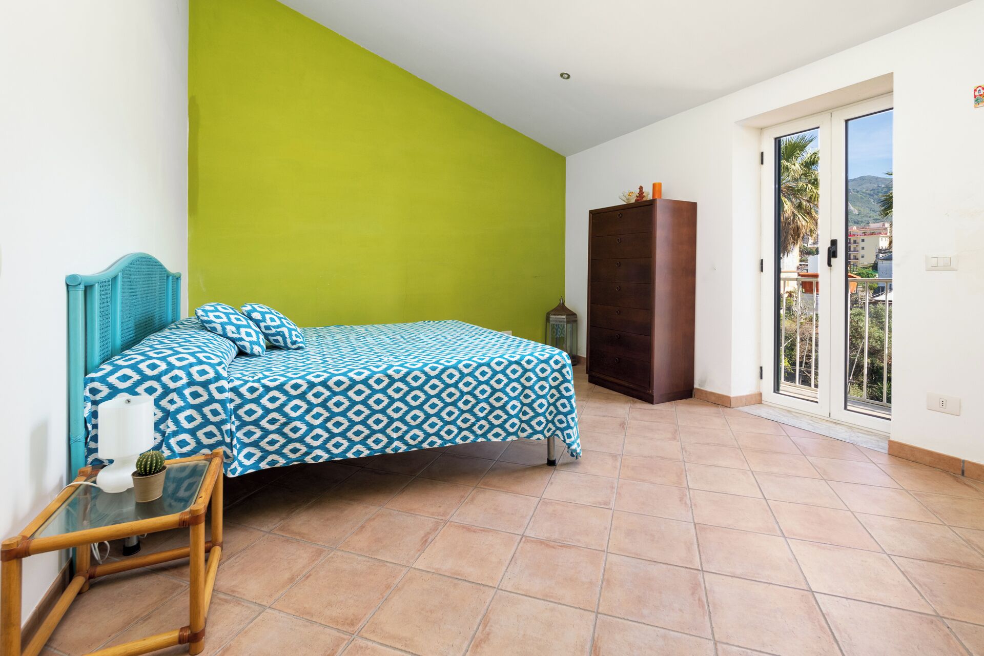 A bedroom with a bed, a dresser, a glass table with a cactus, and a view of the outside through a sliding door.