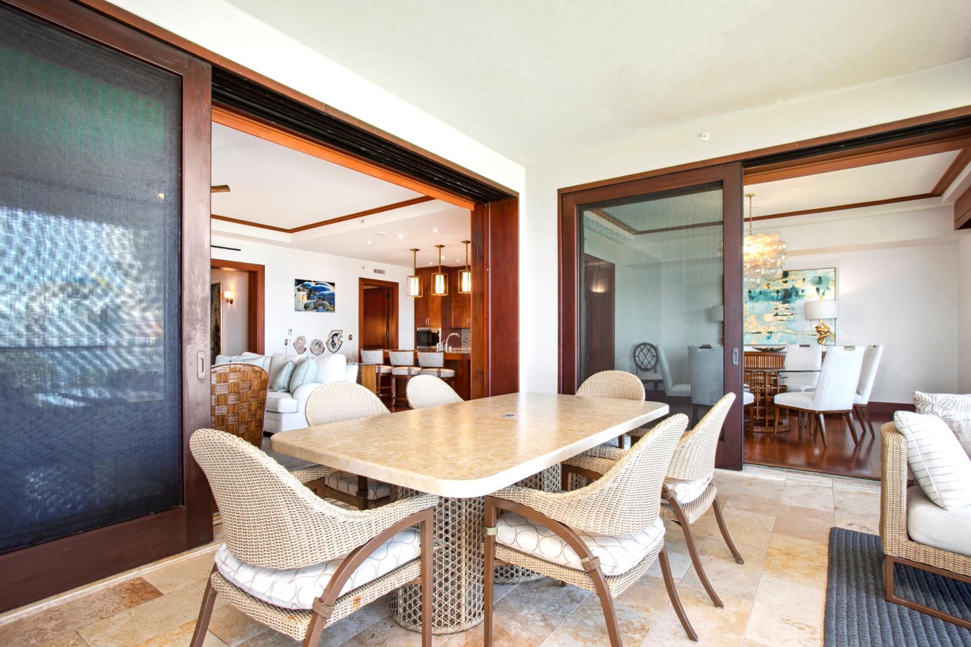 A dining area with a large table and wicker chairs, a kitchen in the background, and a view through a glass door.