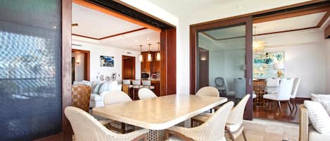 Uma sala de jantar com uma mesa grande e cadeiras de vime, cozinha ao fundo e vista através de uma porta de vidro.