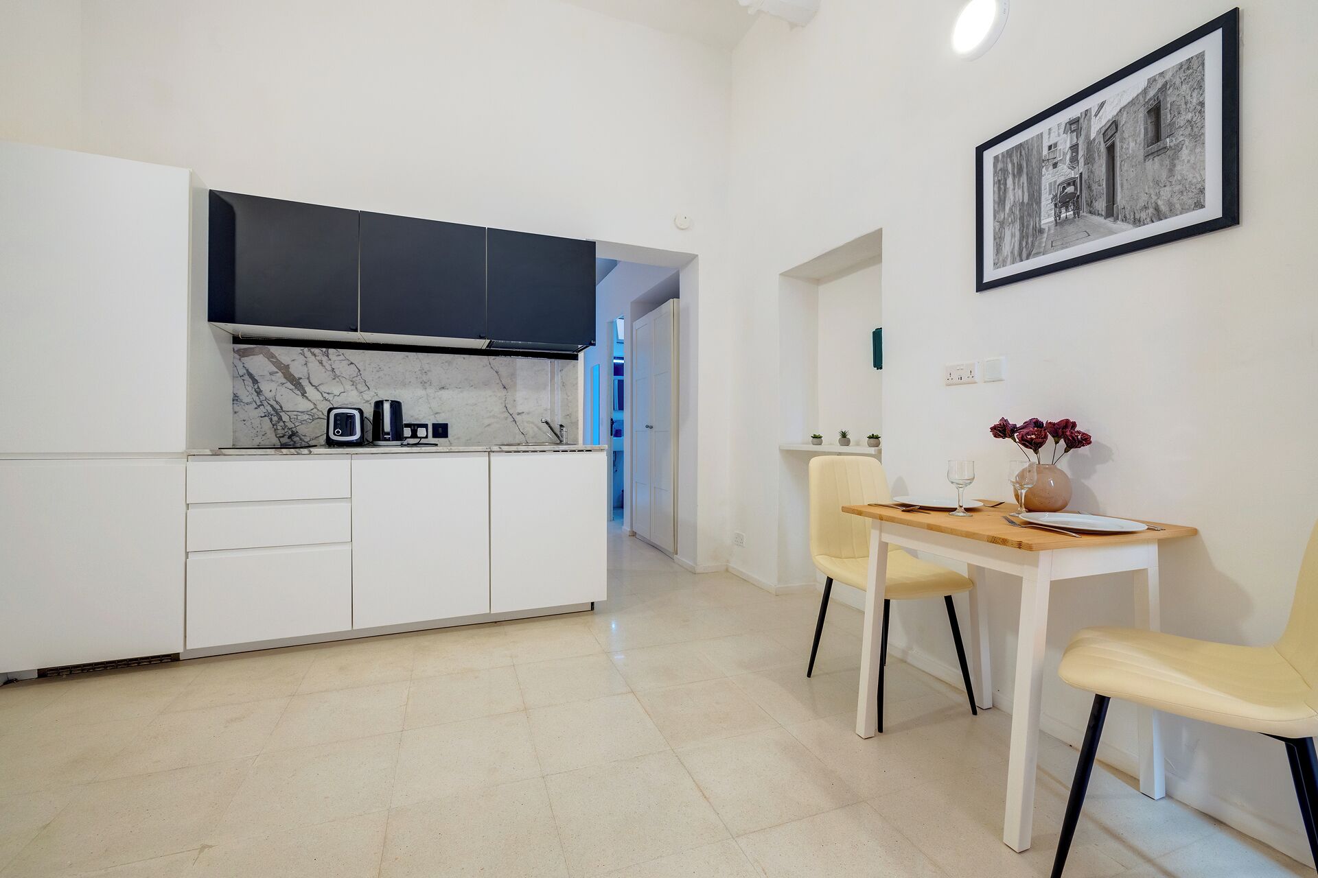 Eine moderne Küche mit weißen Schränken, einer Marmor-Spritzschutzleiste und einer schwarzen Dunstabzugshaube. Es gibt einen Essbereich mit einem Holztisch und zwei Stühlen. An der Wand hängt ein gerahmtes Schwarz-Weiß-Foto.