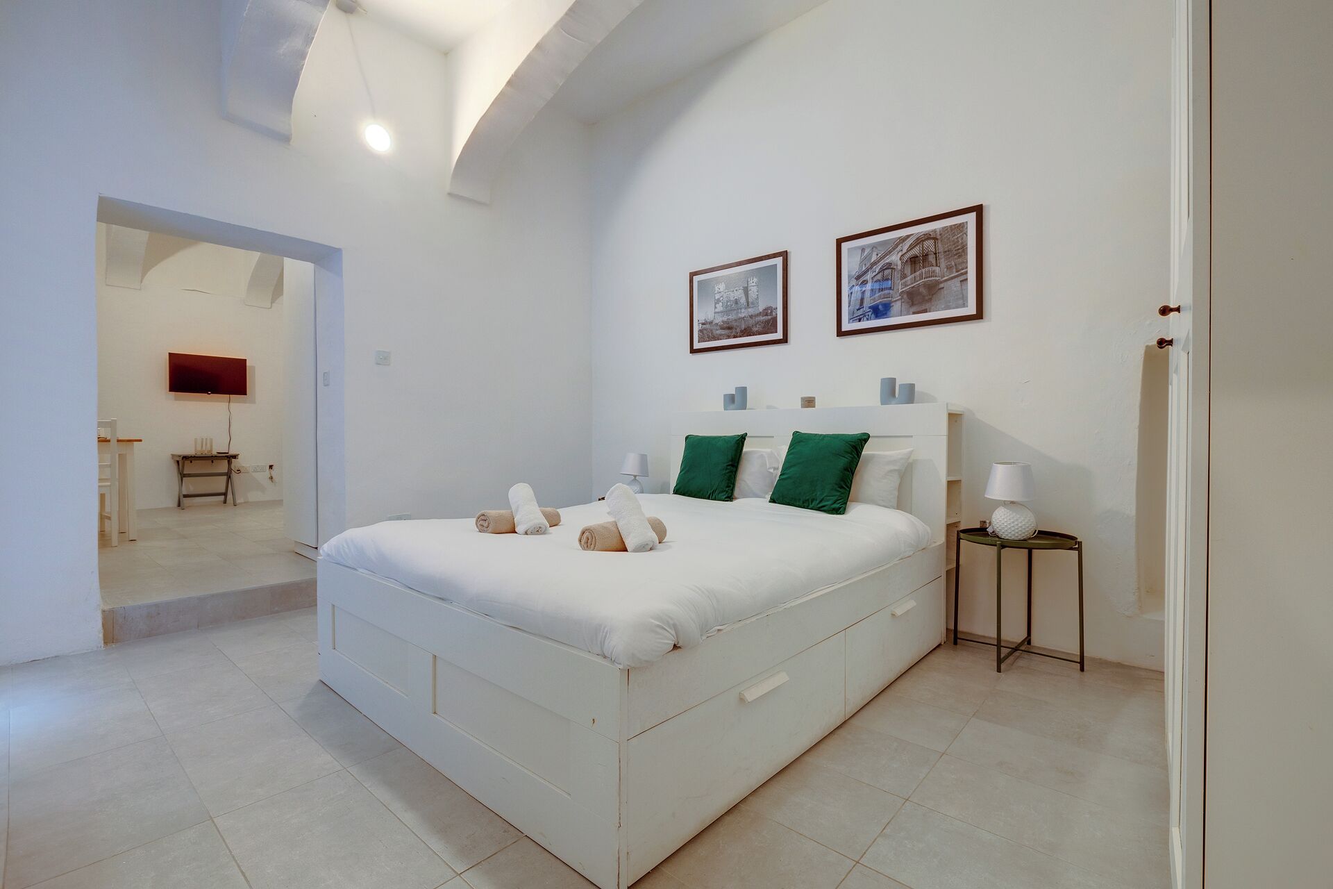 A modern bedroom with a large bed, two green pillows, a bedside table with a lamp, and two framed pictures on the wall.