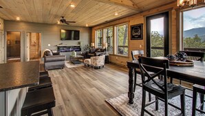 A spacious living area with a wooden ceiling, a fireplace, a dining table, and a large window offering a view of the outdoors.