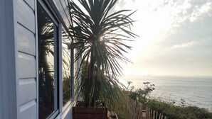 A wooden deck with a view of the ocean, a white building, and a tall plant. - La Bayeuzen - Cabane Balnéo Panoramic sea view - Normandie Arromanches (Saint-Côme-de-Fresné)