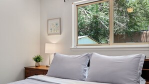 Un dormitorio con cama, mesita de noche con lámpara, cuadro enmarco y ventana con vistas a árboles y un edificio.