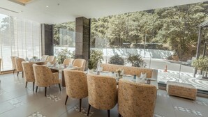 A restaurant with a view of a forested area, featuring a long table with floral chairs and a small round table with a vase. - NAMAMI BY ZENQ BANKHANDI (Dera Gopipur)