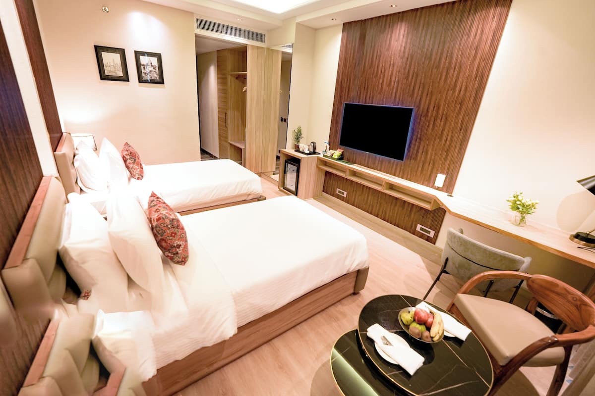 Quarto de hotel com duas camas, uma televisão, uma escrivaninha com cadeira e uma mesinha com fruta.
