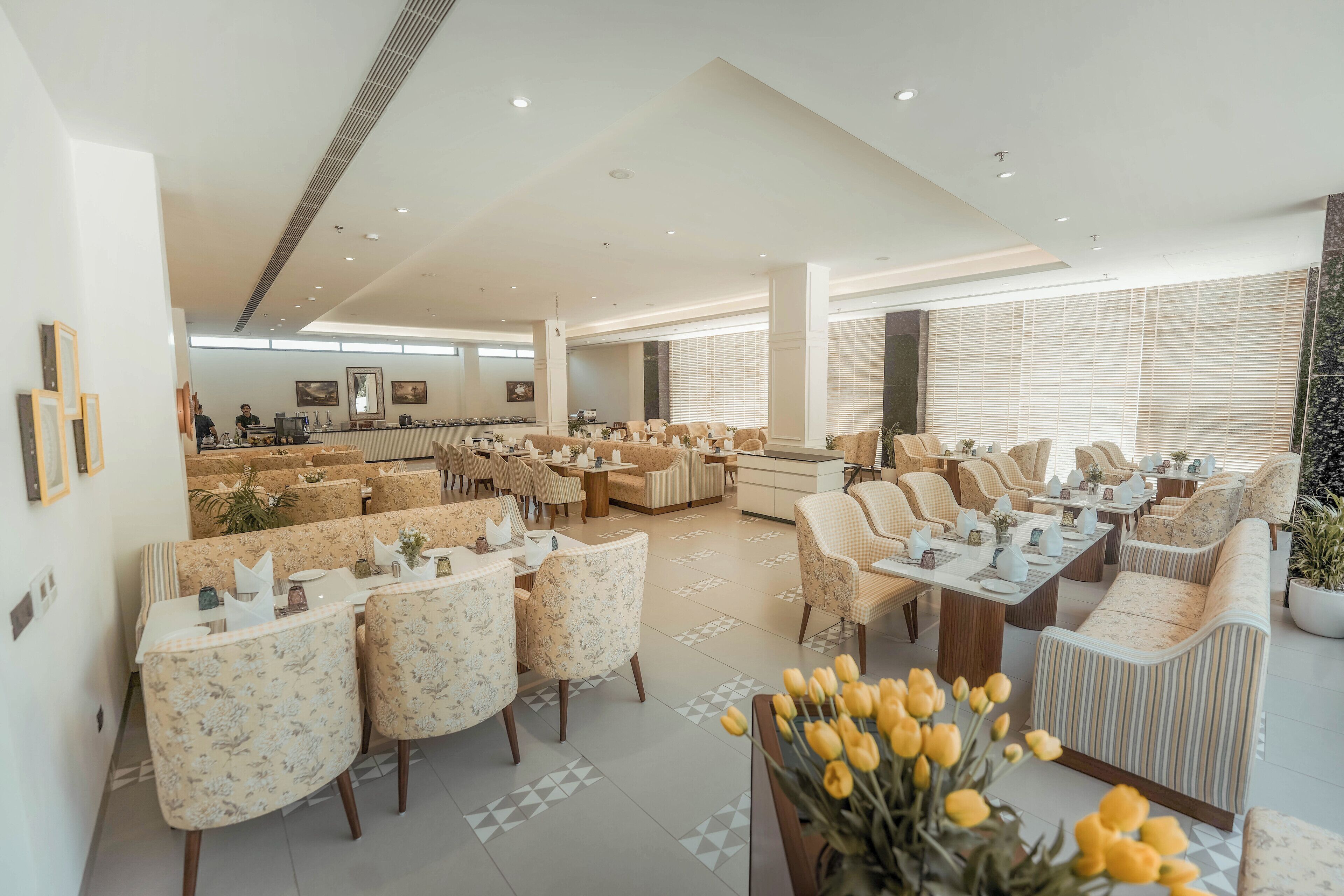 A spacious hotel lobby with multiple seating areas, including a dining area with tables and chairs, and a lounge with sofas and armchairs.
