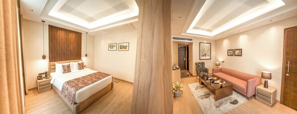 A hotel room with a large bed, a sofa, a coffee table, and a desk. There are framed pictures on the walls and a plant on the floor.