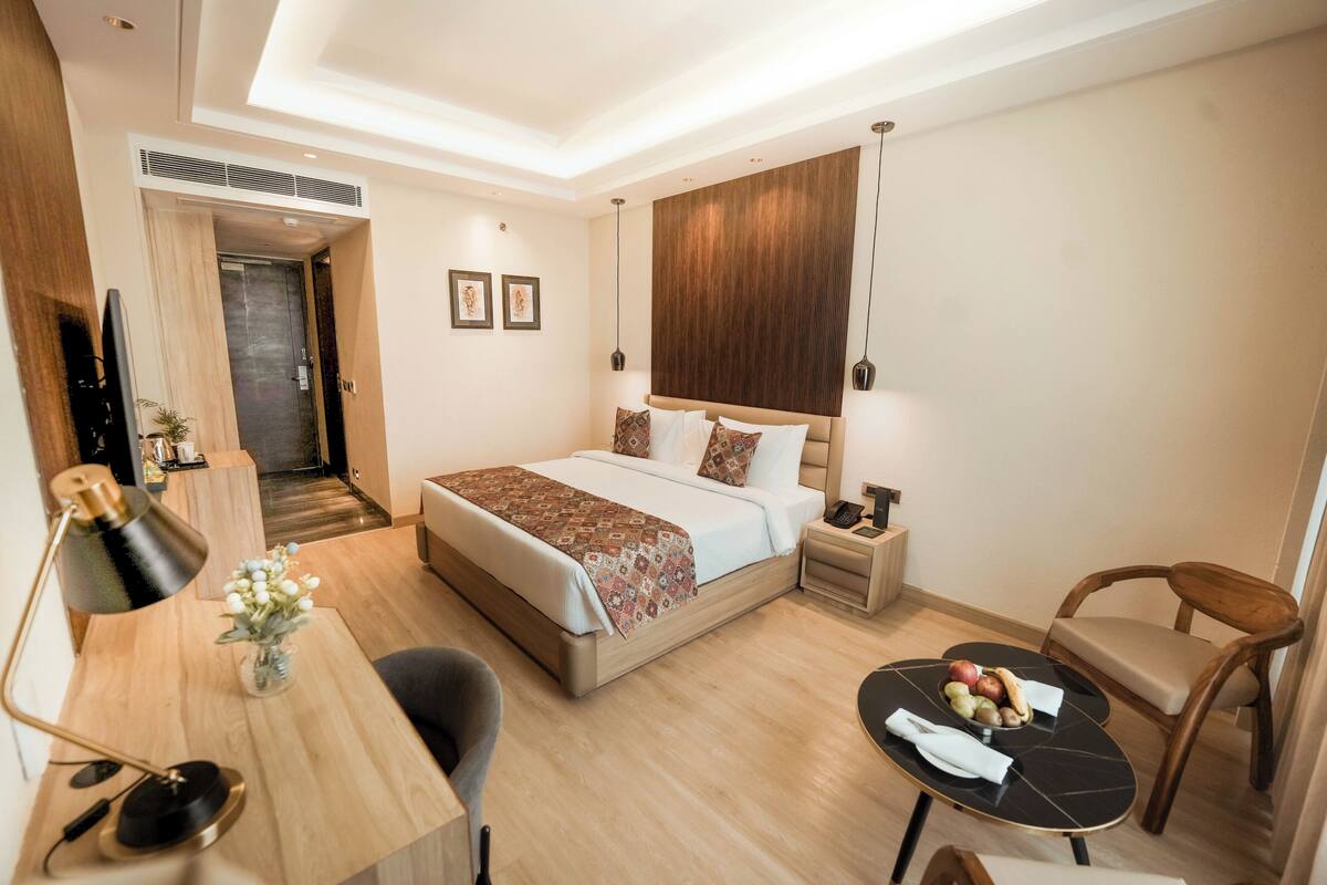 Quarto de hotel moderno com cama, mesa de cabeceira, escrivaninha, cadeira e uma pequena mesa de jantar com frutas.