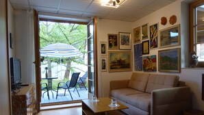 Un salon avec un canapé, une table basse et un téléviseur. Dehors, sous un parasol, on peut voir des chaises.