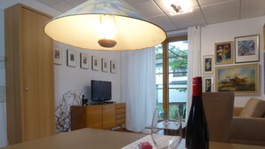 Un coin repas avec une table dressée pour deux personnes, une bouteille de vin et un verre. On y trouve également un téléviseur posé sur un meuble en bois, une fenêtre donnant sur des arbres et des œuvres d’art encadrées accrochées au mur.
