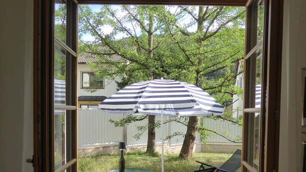 Une porte ouverte donne sur une terrasse où se trouvent une table et des chaises, un parasol à rayures, et d’où l’on peut admirer un jardin avec des arbres et une clôture.