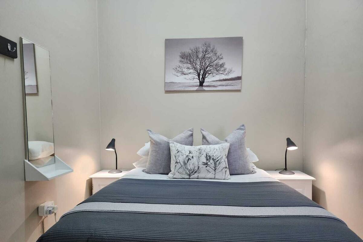 A bedroom with a bed, two bedside tables, a night lamp, a wall-mounted mirror, and a framed picture of a tree.