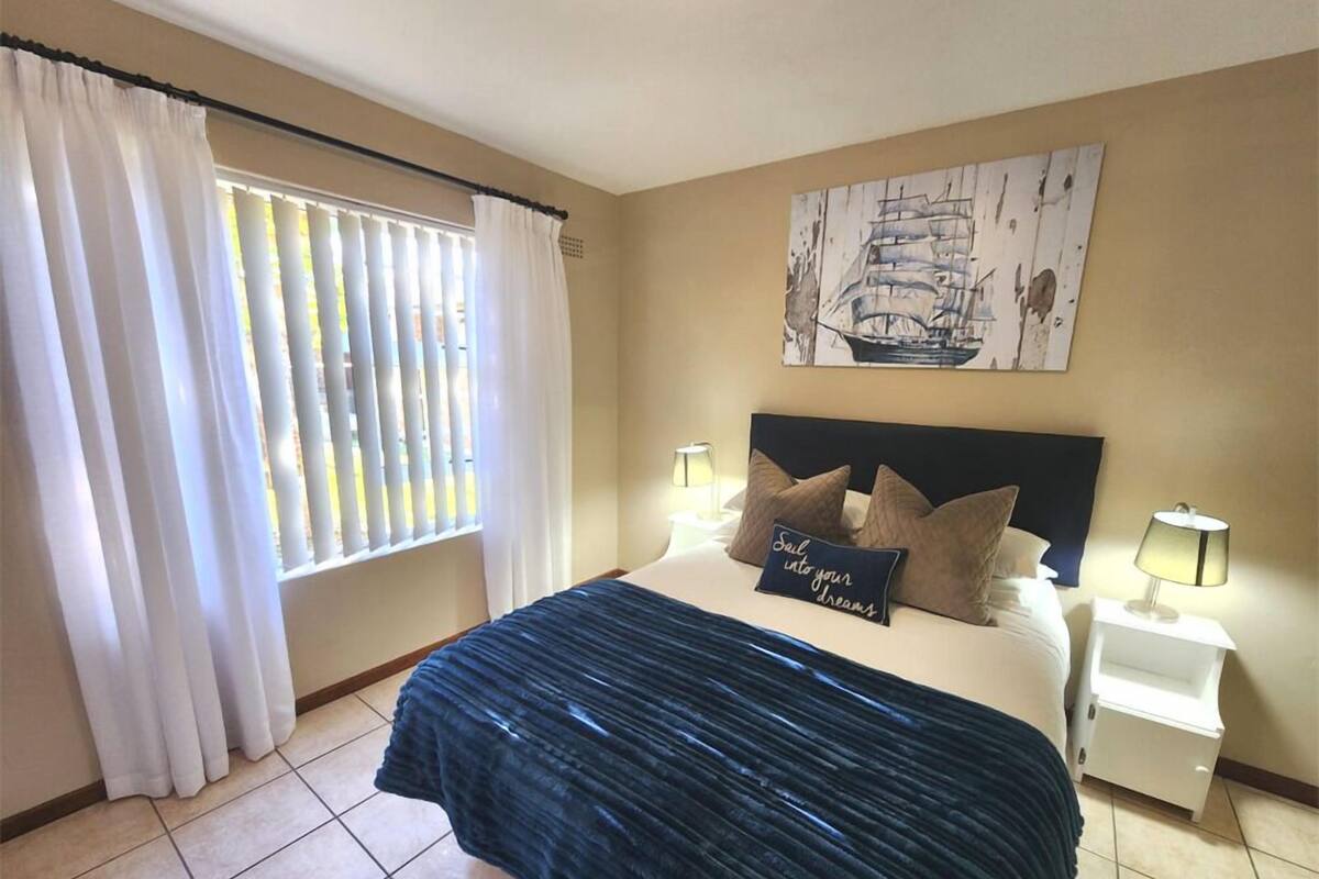 A bedroom with a bed, a nightstand, a lamp, a window with curtains, and a wall art piece.