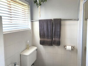 A bathroom with white tiles, a toilet, a towel rack with two towels, a window with blinds, and a hanging plant. - Muir Holiday Apartments (Still Bay)