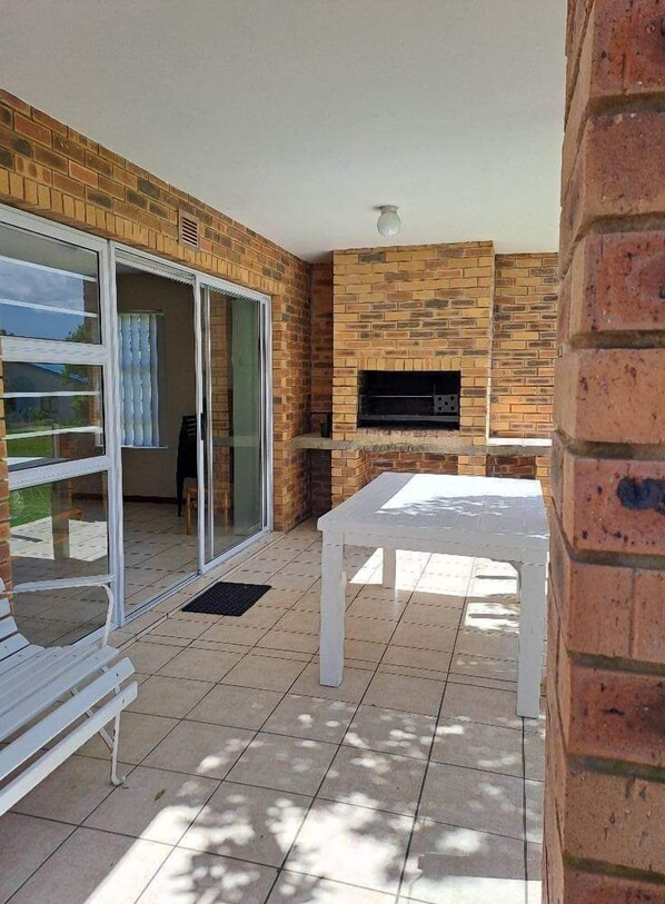 An outdoor patio with a brick wall, a sliding glass door, a white table, and a flat-screen TV mounted on the wall. - Muir Holiday Apartments (Still Bay)