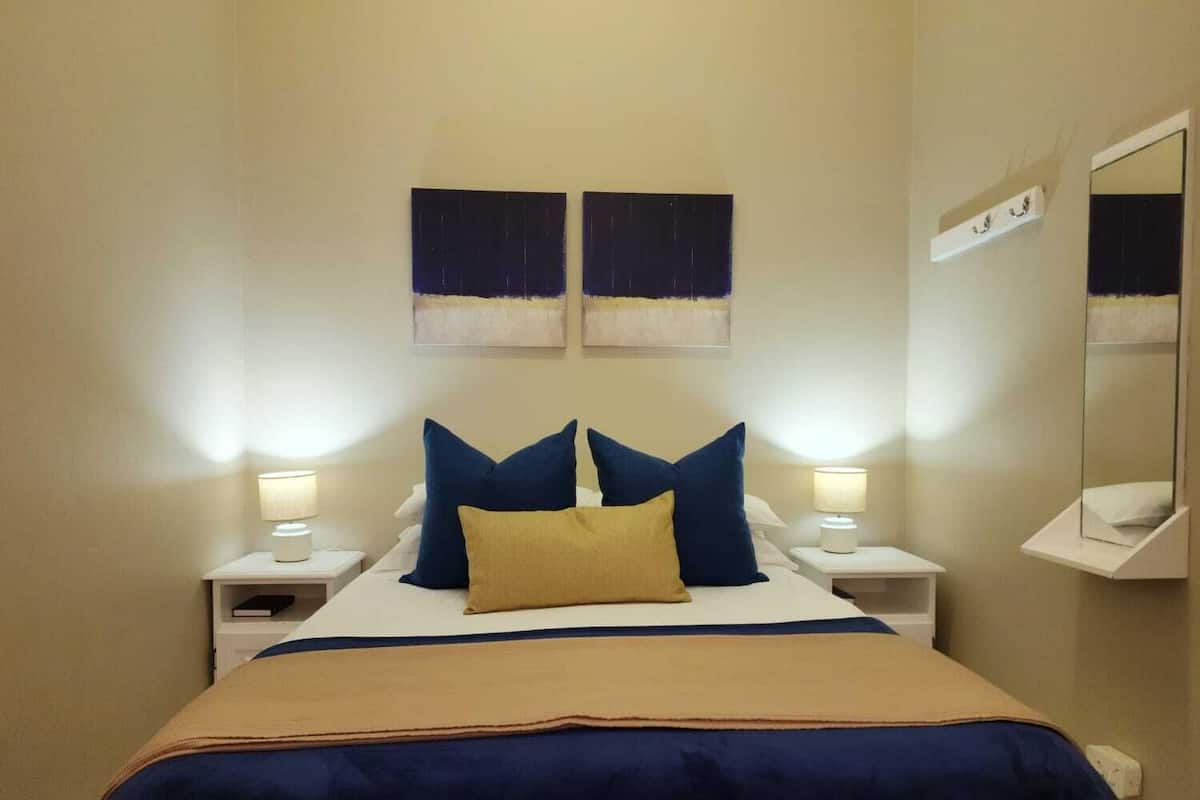 A hotel room with a bed, two bedside lamps, a nightstand, a mirror, and two wall-mounted paintings.