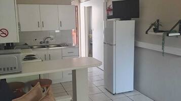 A compact kitchen with white cabinets, a refrigerator, a microwave, and a television mounted on the wall.