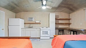 Eine kleine Küche mit Kühlschrank, Herd, Mikrowelle und Holzregalen. Zwei Betten mit orange- und blau gestreuter Bettwäsche.