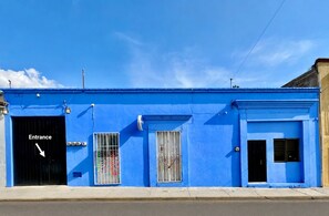 A blue building with a black entrance door, a window with a metal gate, and a small window with a curtain.