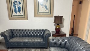 A living room with a dark grey tufted sofa, a wooden coffee table, and a lamp. There are framed botanical prints on the wall. - AggieLand Vacation House!  Charming 3-BR 2-BA only 3 miles from Texas A&M Campus (Bryan)