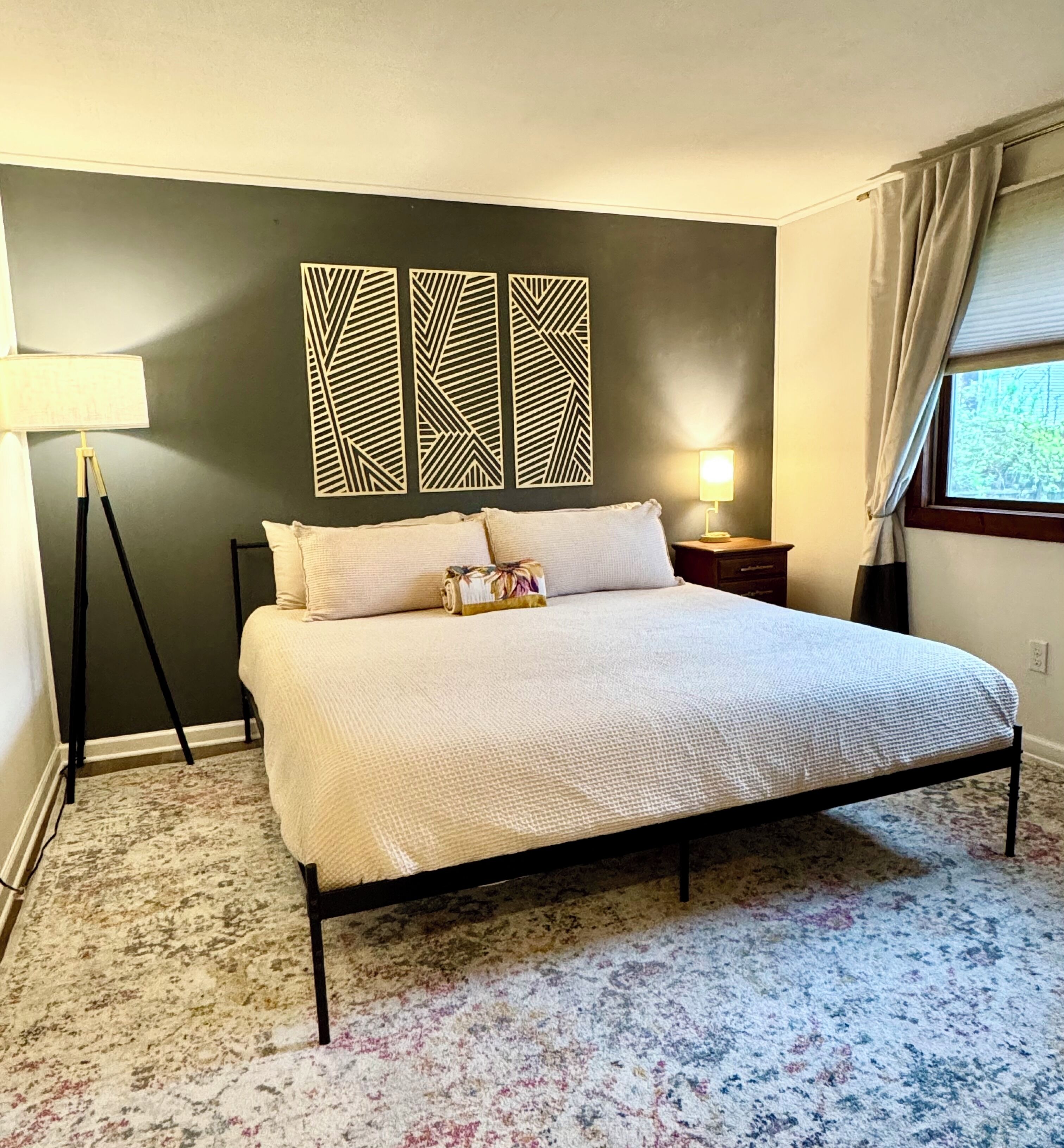 A bedroom with a bed, a lamp, a window with curtains, and a wall with geometric patterns.