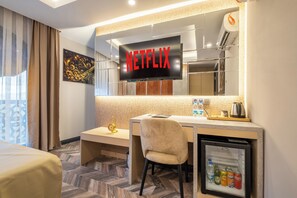 A hotel room with a minibar, a desk, a chair, a bed, a television displaying 'NETFLIX', and a small refrigerator.