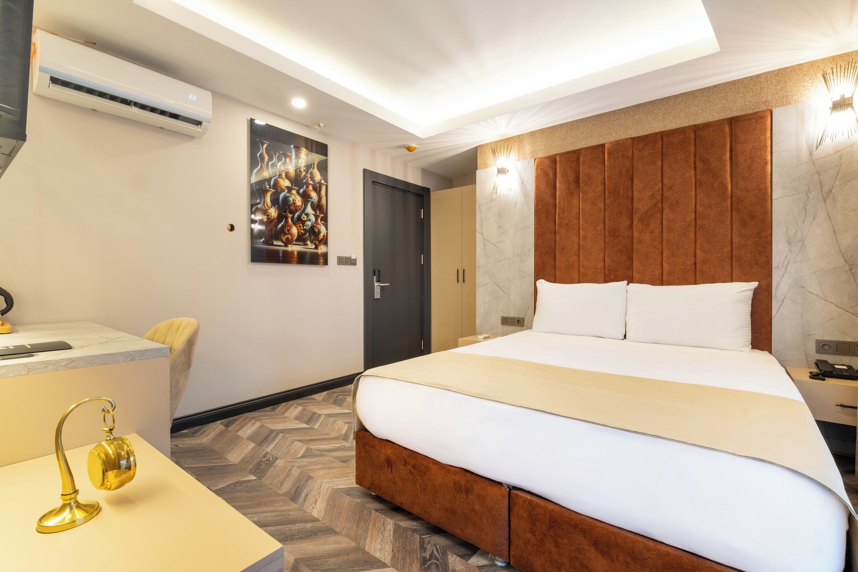 A modern hotel room with a large bed, a desk with a lamp, and a herringbone-patterned floor.