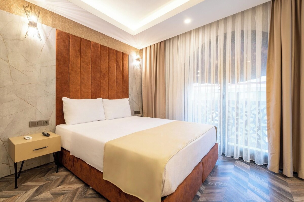 A hotel room with a large bed, a nightstand, a wooden headboard, and a view of the outdoors through sheer curtains.