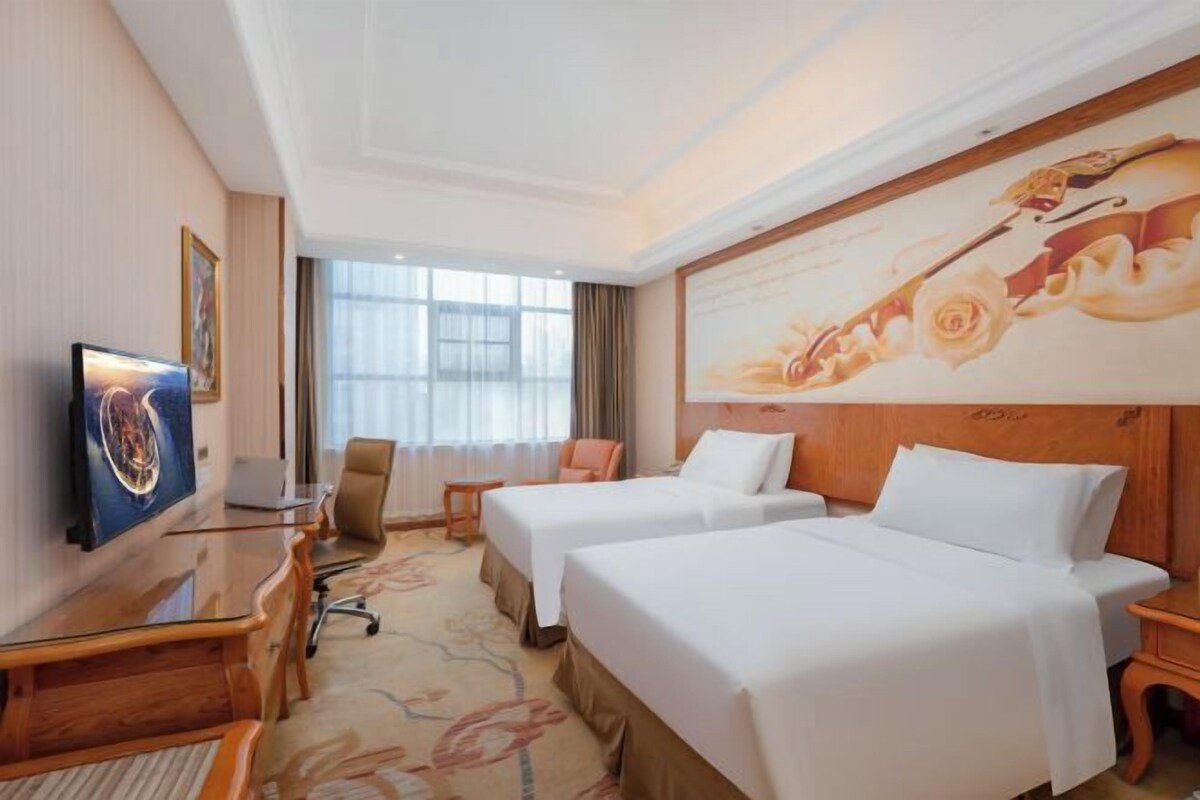A hotel room with two beds, a desk, a chair, and a large window.