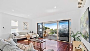 A modern living room with a grey sofa, a coffee table, and a balcony with outdoor furniture. - Manly Oasis 5BR - 3 King Beds, Pool, BBQ (Manly)