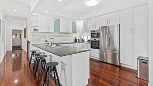 A modern kitchen with white cabinetry, a central island with stools, stainless steel appliances, and a wooden floor. - Manly Oasis 5BR - 3 King Beds, Pool, BBQ (Manly)
