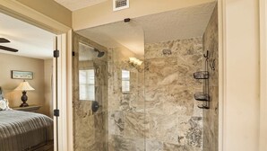 Une douche à l’italienne avec des carreaux de marbre et une porte en verre, un lit avec une couette beige et une table de chevet avec une lampe.