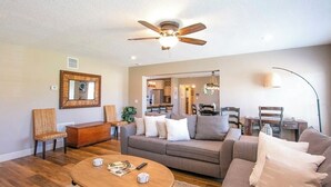 Un salon spacieux doté d’un ventilateur de plafond, d’un grand canapé, d’une table basse et d’un coin repas visible à travers une porte ouverte.