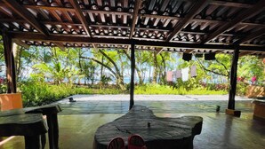 Un espace extérieur abrité, doté d’un banc en bois, d’une table et offrant une vue sur un jardin luxuriant et une plage.