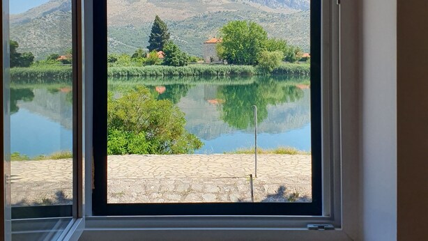 A room with a large window overlooking a lake, mountains, and greenery.