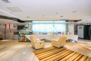 Zona de recepción de un hotel con mostrador, dos sillones, un tapete estampado y una mesa con varios objetos.