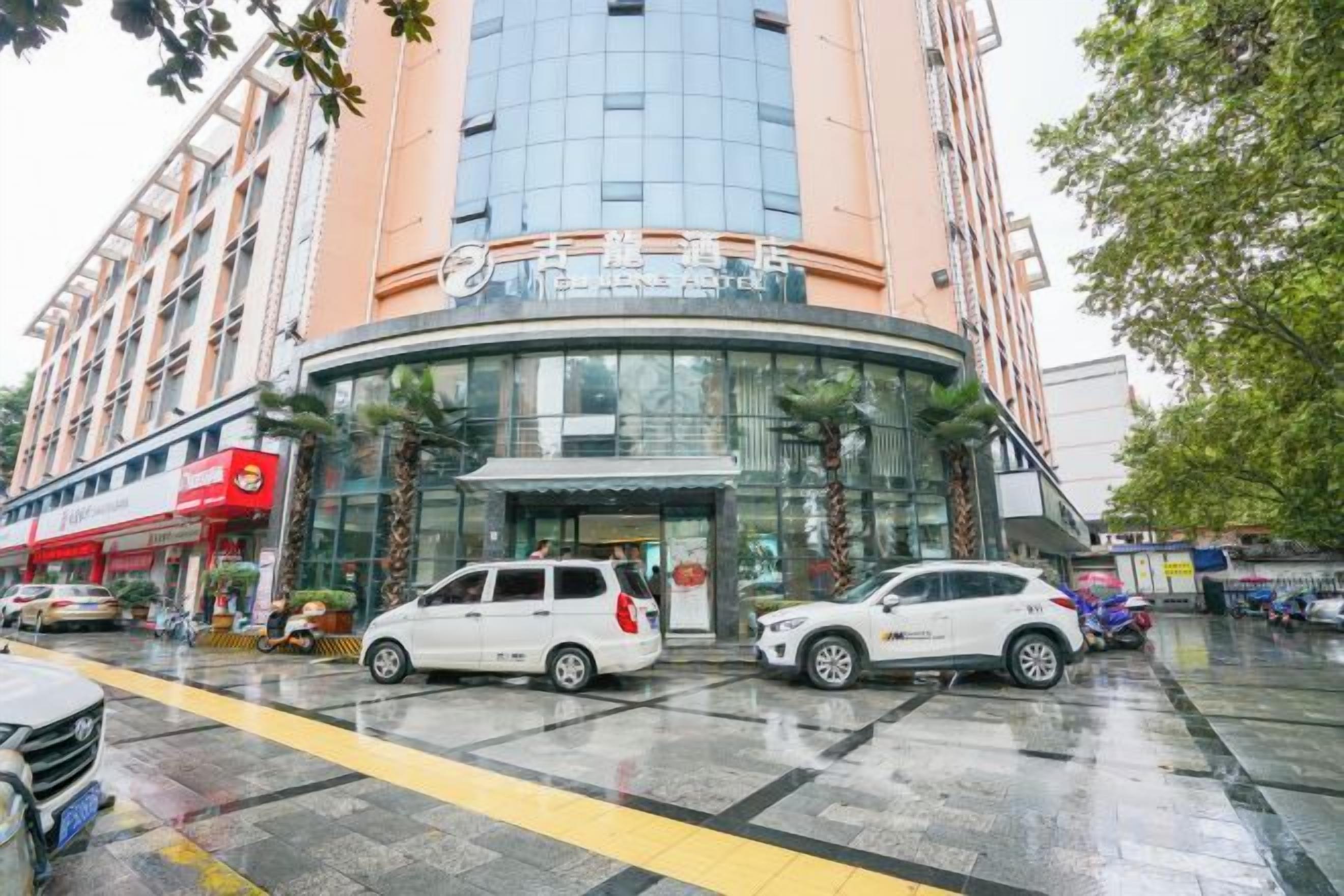 A multi-story building with a curved glass facade, a parking area with cars, and palm trees.