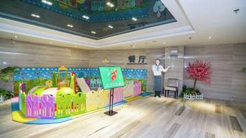 A children's play area with a slide and climbing structures, a reception desk, and a decorative wall with a cherry blossom tree.