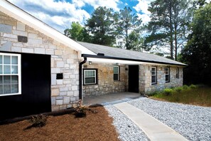 A stone house with a black door and windows, surrounded by trees and a gravel driveway. - Modern 4BR Home - Very Spacious - Minutes from Georgia International Horse Park (Conyers)