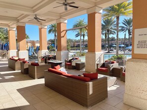 An outdoor lounge area with wicker furniture and red cushions, a pool in the background, and a ceiling fan.