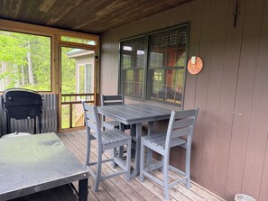 A covered outdoor seating area with a table and chairs, a wooden deck, and a view of the surrounding greenery.